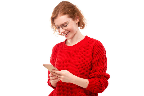 Gelukkig glimlachende roodharige meisje houden smartphone in handen in bril en rode kleren geïsoleerd op witte studio achtergrond, kreeg een vreugdevolle boodschap - Foto, afbeelding
