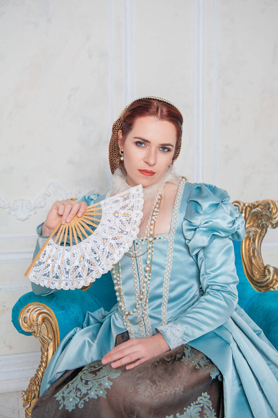 Beautiful young woman in medieval style blue dress with fan sitting in the armchai - Photo, Image