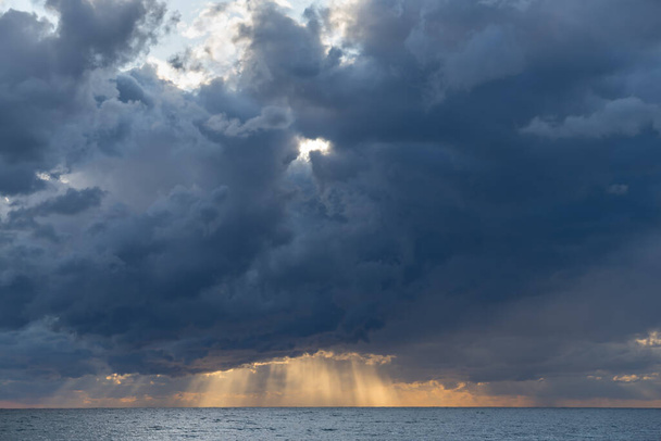 Diep donkerblauwe wolken met zeldzame zonnevlekken daalden laag over de Zwarte Zee. De gele stralen van de zon zijn zichtbaar en verlichten het water. De textuur van de stormachtige lucht. Volumetrische natuurfoto  - Foto, afbeelding