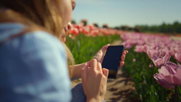 Задний вид молодой женщины, касающейся экрана смартфона пальцами в цветочном поле - Фото, изображение