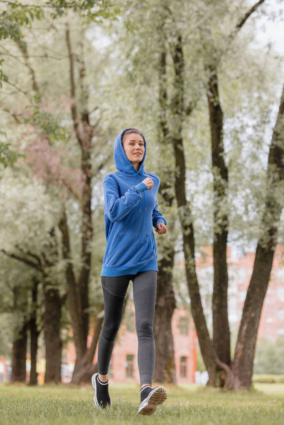 a European woman in a blue hoodie is running and doing sports in the park. fitness and yoga outdoor sports - Fotografie, Obrázek