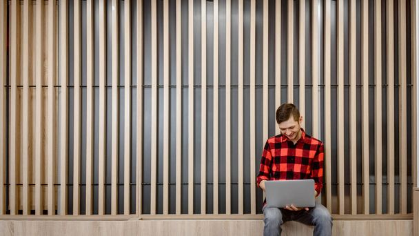 Ευτυχισμένος νεαρός άνδρας σε casual καρό πουκάμισο προγραμματιστής λογισμικού εργάζεται σε ένα ενδιαφέρον γραφείο με ξύλο πίνακες τοίχου στο laptop του - Φωτογραφία, εικόνα
