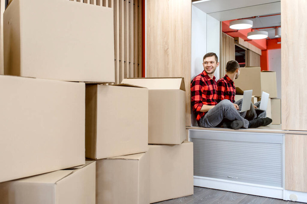 Счастливый улыбающийся молодой человек, предприниматель, бизнесмен, переехал в новый офис и работает над своим ноутбуком рядом с большим количеством картонных коробок. Концепция движения - Фото, изображение