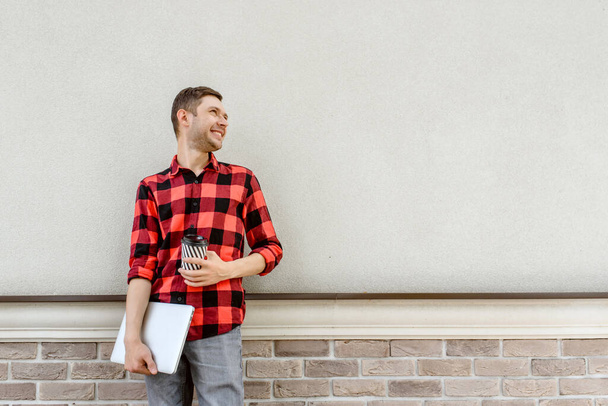 Joven hombre feliz freelancer está de pie cerca de una pared de ladrillo y la celebración de un café y un ordenador portátil en sus manos. Trabajando en todas partes concepto. Espacio de copia para tex - Foto, imagen