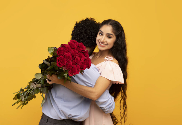 Felice signora indiana abbracciando il suo ragazzo e tenendo bouquet di rose, celebrando San Valentino su sfondo giallo - Foto, immagini