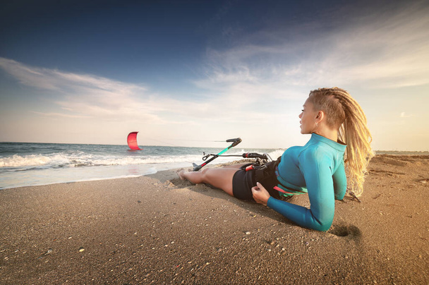 Attractive caucasian woman with dreadlocks on her head in a wetsuit lies on a sandy beach and holds her kite. Water sports. Kite surfer on vacation. Copy space - Photo, Image
