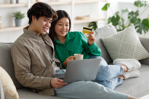 Happy Asian couple shopping online via laptop and credit card, ordering things in internet, sitting on sofa at home - Photo, Image