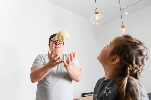 Having fun. Man in glasses throwing up dough while feeling playful with his caucasian little daughter, while preparing homemade cookies. Girl looking at the father - Foto, Imagem