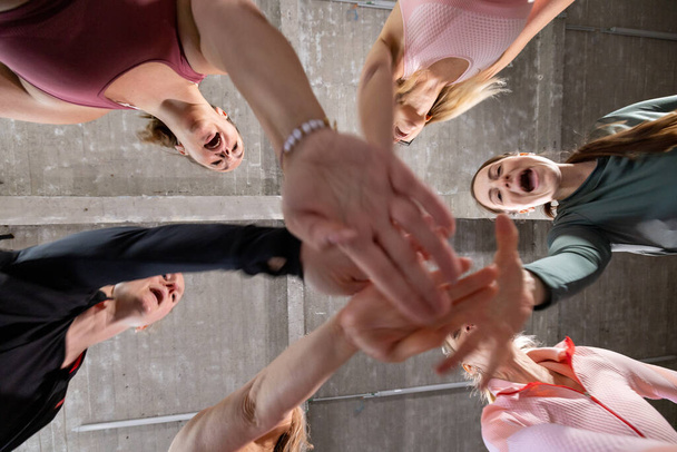 Grupa sześciu młodych zmotywowanych dziewcząt sportowych noszących strój sportowy stojących razem tworząc stos dłoni przed treningiem w klasie fitness. - Zdjęcie, obraz