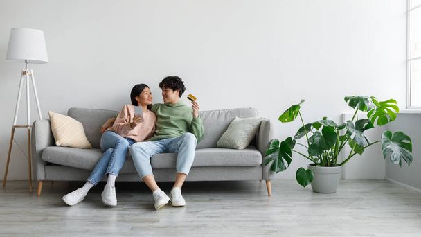 Cheerful young Asian couple sitting on sofa with credit card and cellphone, shopping online in living room, copy space - Φωτογραφία, εικόνα