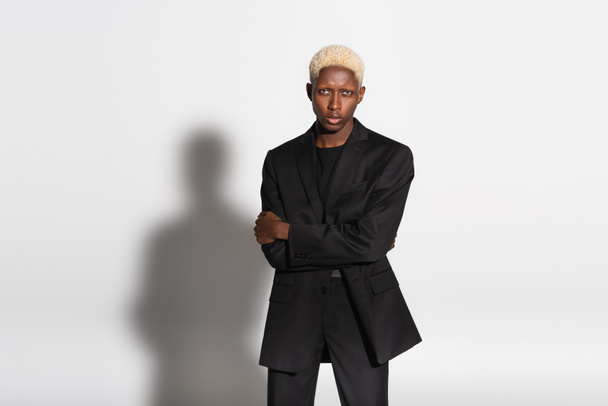 blonde african american man in black suit standing with crossed arms on grey with shadow - Zdjęcie, obraz