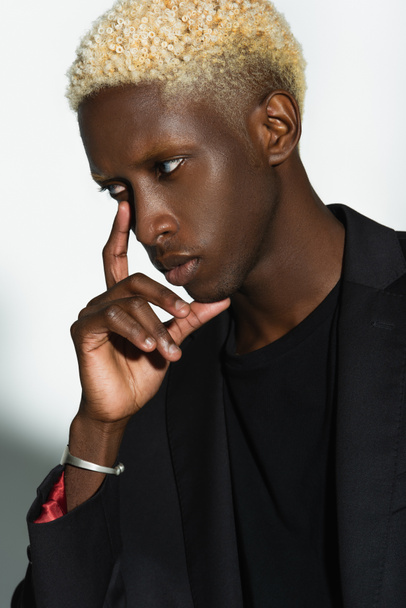 young african american man with blonde hair posing with hand near face on grey - Fotó, kép