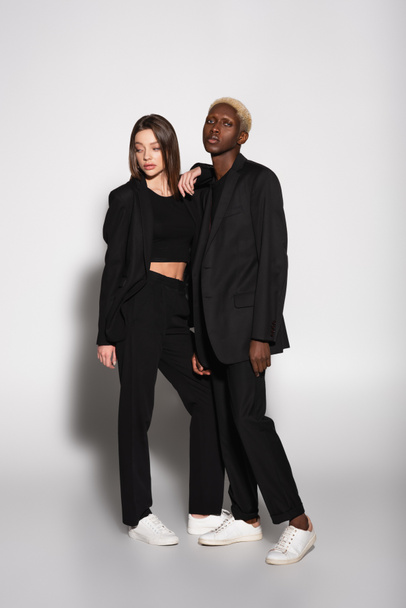 μοντέρνο πολυεθνικό ζευγάρι με μαύρα ρούχα και λευκά sneakers που ποζάρουν στο γκρι με σκιά - Φωτογραφία, εικόνα