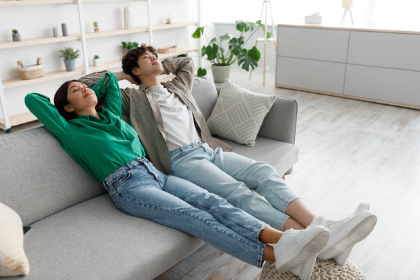 Calm young Asian couple relaxing on couch with their hands behind heads at home, blank space - Фото, изображение