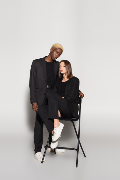 blonde african american man looking at camera near brunette woman on chair on grey with shadow - Photo, Image