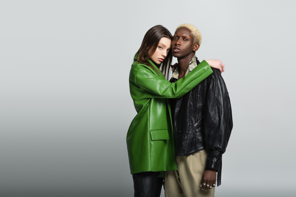 pretty woman in green leather jacket hugging stylish african american man on grey - Photo, Image