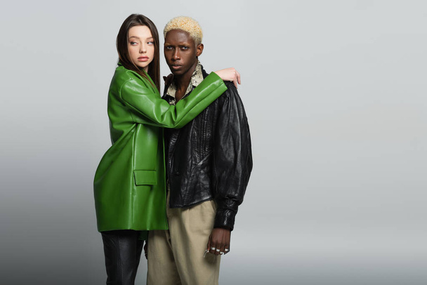stylish woman in green leather jacket embracing blonde african american man looking at camera on grey - Foto, immagini