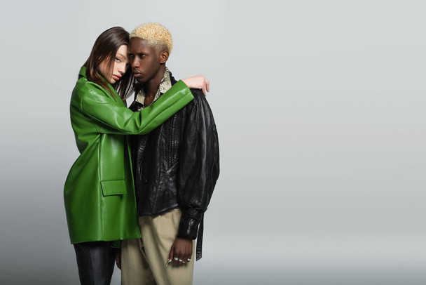 mujer joven en chaqueta de cuero verde abrazando al hombre afroamericano rubio y mirando la cámara en gris - Foto, Imagen