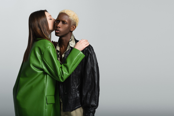 brunette woman in stylish jacket kissing african american man with bleached hair isolated on grey - Fotó, kép