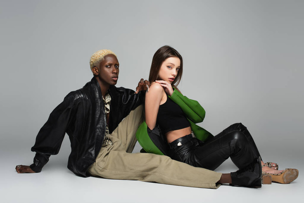 blonde african ameican man and sensual brunette woman sitting and looking at camera on grey - Photo, Image