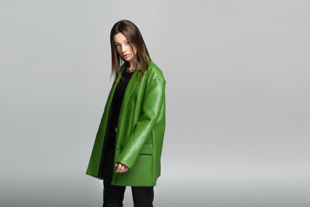 young and stylish woman in green leather jacket looking at camera isolated on grey - Photo, Image