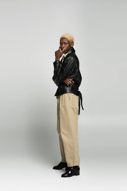 full length view of blonde african american man in black leather jacket and beige trousers on grey - Fotografie, Obrázek