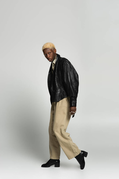 full length view of blonde african american man in beige trousers and black jacket posing on grey - 写真・画像