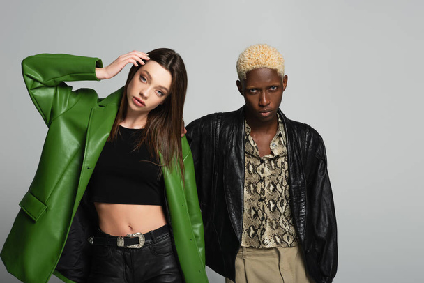 young woman in black top and green leather jacket posing near blonde african american man isolated on grey - Photo, Image