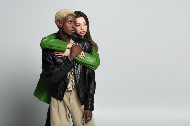 brunette woman embracing blonde african american man in trendy clothes and looking away on grey - Фото, изображение