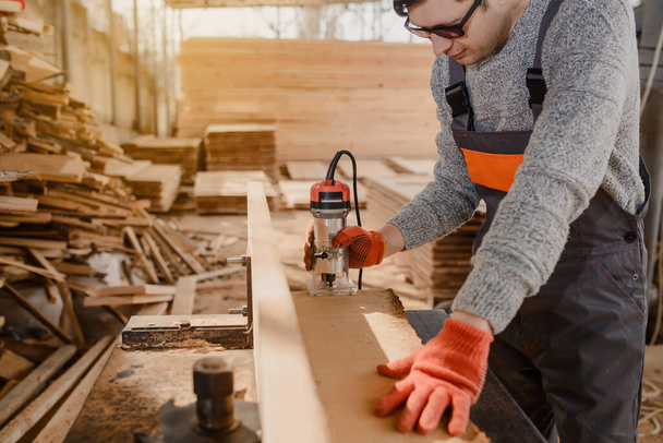 Jeune homme charpentier à l'entrepôt de l'atelier en bois utilise une fraiseuse sur une planche / planche en bois à partir d'une grande pile de bois de planches en bois. - Photo, image