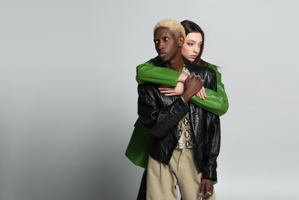 young woman hugging blonde african american man in black leather jacket on grey - Photo, Image