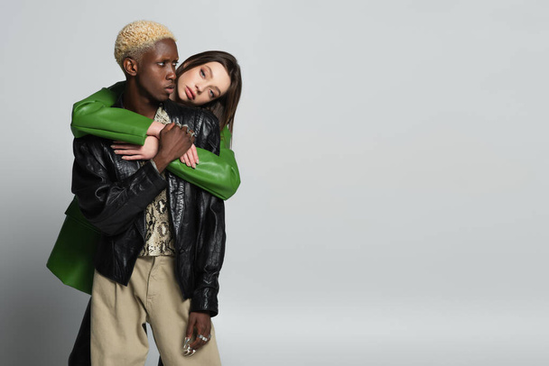 young woman looking at camera while hugging stylish african american man with bleached hair on grey - Foto, Imagen