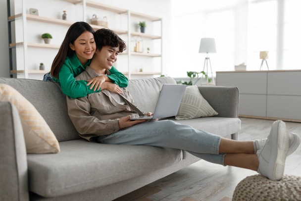 Junge Asiatin umarmt ihren Freund, benutzt Laptop, surft im Internet auf der heimischen Couch, leerer Raum - Foto, Bild