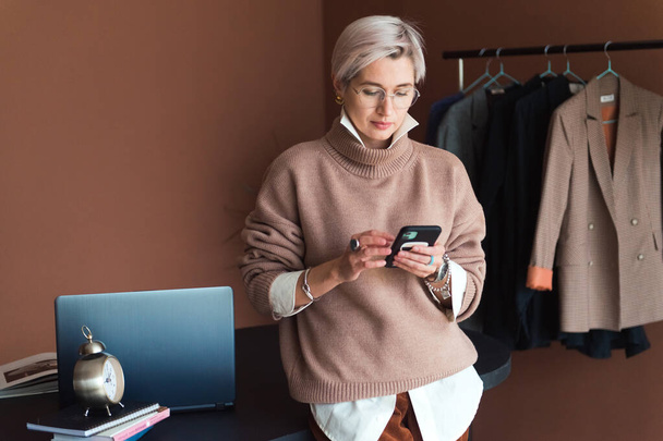 fashion woman in brown oversize sweater and trousers standing in modern work place or office with smartphone - Valokuva, kuva