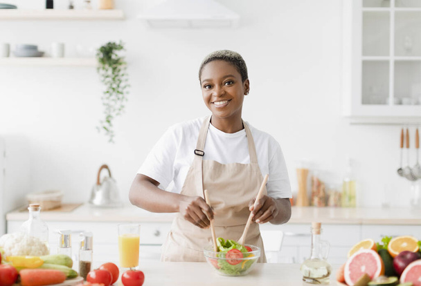 Usmívající se tisíciletá hezká černoška v zástěře dělá salát v kuchyni interiér u stolu s organickým ovocem - Fotografie, Obrázek