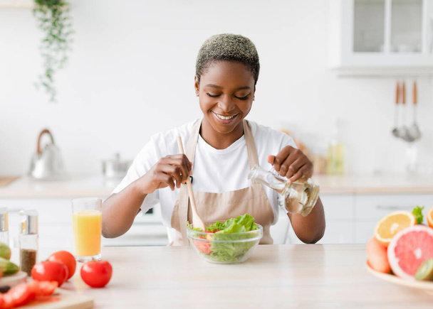 Soddisfatto giovane bella signora nera in grembiule rende insalata a cucina interna a tavola con frutta biologica - Foto, immagini