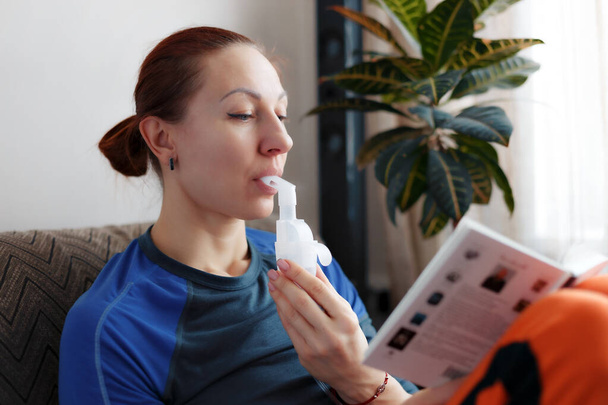 Mulher branca está lendo livro e segurando tubo de nebulizador, inalando vapores de medicamentos em seus pulmões. - Foto, Imagem