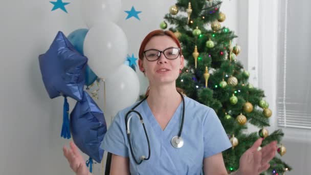 fiatal női orvos szemüveges gratulál a családjának videohívással, miközben ül az irodában karácsonyfa, miközben dolgozik az újévi ünnepek - Felvétel, videó