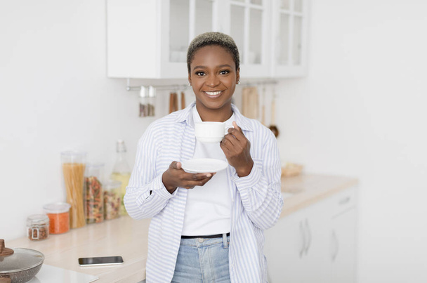Веселый довольно тысячелетняя черная женщина в повседневной одежде пить свой любимый горячий напиток в белой чашке - Фото, изображение