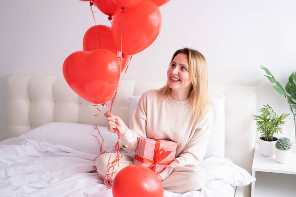 Beautiful women in casual cozy clothes on bed with red heart balloons, celebrating Valentine day - Fotó, kép