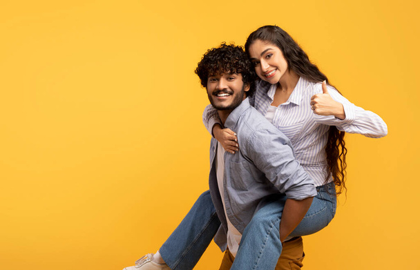 Gran oferta. Emocionado indio hombre piggybacking novia y mujer mostrando pulgar hacia arriba, fondo amarillo, espacio de copia - Foto, Imagen