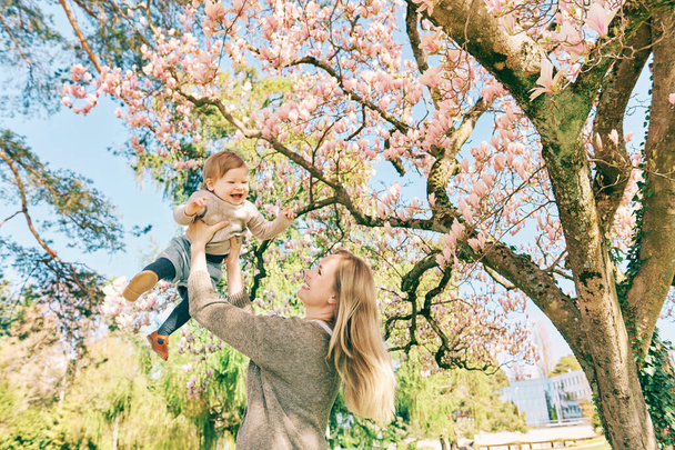 Εξωτερική πορτρέτο της ευτυχισμένης νεαρής μητέρας με αξιολάτρευτο κοριτσάκι κάτω από ανθισμένο δέντρο άνοιξη, έχοντας καλές στιγμές στο ηλιόλουστο πάρκο - Φωτογραφία, εικόνα