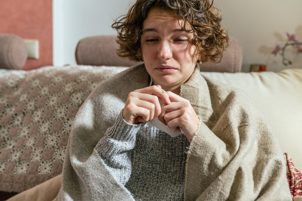 Concept of seasonal cold and weakened immune system. Sick young woman home. Upset young woman with runny nose and flu or allergy symptoms is about to sneeze. girl with disposable napkin is wrapped in blanket home on couch. - Photo, Image