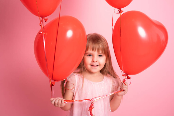 χαριτωμένο κορίτσι παιδί σε ροζ φόρεμα με κόκκινα μπαλόνια καρδιά σε ροζ φόντο. Ημέρα του Αγίου Βαλεντίνου - Φωτογραφία, εικόνα