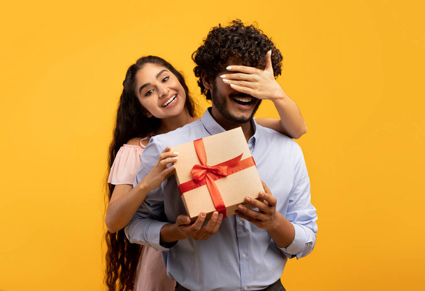 Досить індійська жінка, яка прикриває очі своїм хлопцям, тримає подарункову коробку і вітає його з днем народження або річницею
 - Фото, зображення