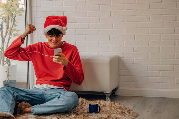 έφηβος αγόρι στο σπίτι τα Χριστούγεννα ενθουσιασμένος κοιτάζοντας το κινητό τηλέφωνο - Φωτογραφία, εικόνα