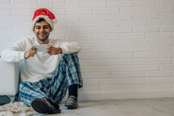 mężczyzna na Boże Narodzenie z kapeluszem Świętego Mikołaja i telefonem komórkowym w domu uśmiechnięty - Zdjęcie, obraz
