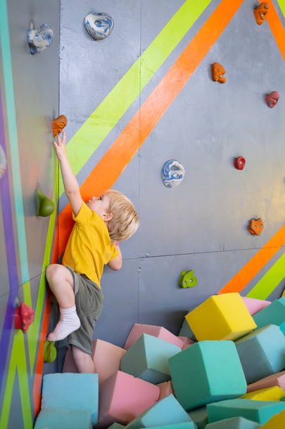 Little cute boy is climbing on vertical wall indoor in entertainment park - Foto, Imagem
