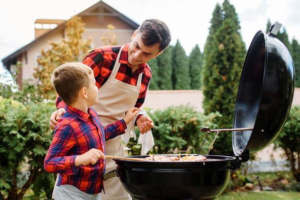 El joven padre y su hijo están cocinando barbacoa en una parrilla al aire libre en el patio trasero de su casa y divirtiéndose. Concepto de familia feliz - Foto, Imagen