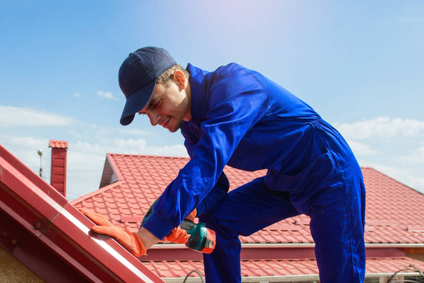 Jonge man werknemer in blauw over het algemeen vast te stellen een metalen tegel dak met schroevendraaier. Begrip dakbedekking - Foto, afbeelding
